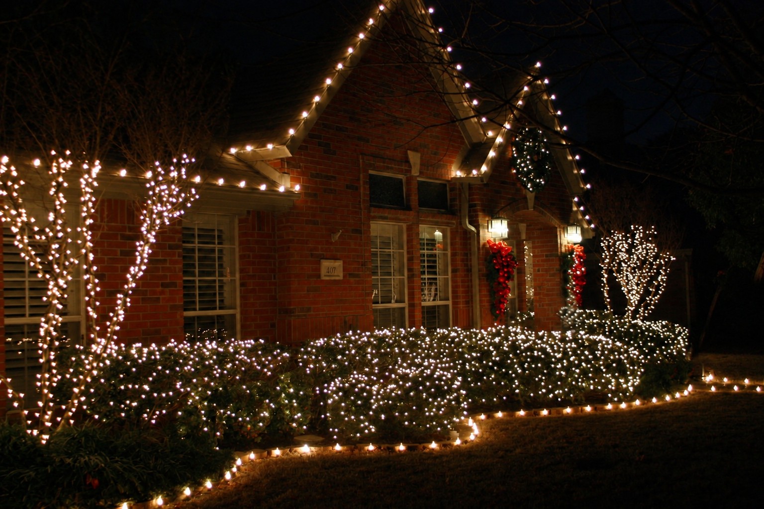 House roofline lighting example White Christmas lights outline a brick house roofline, shrubs and garden edge.